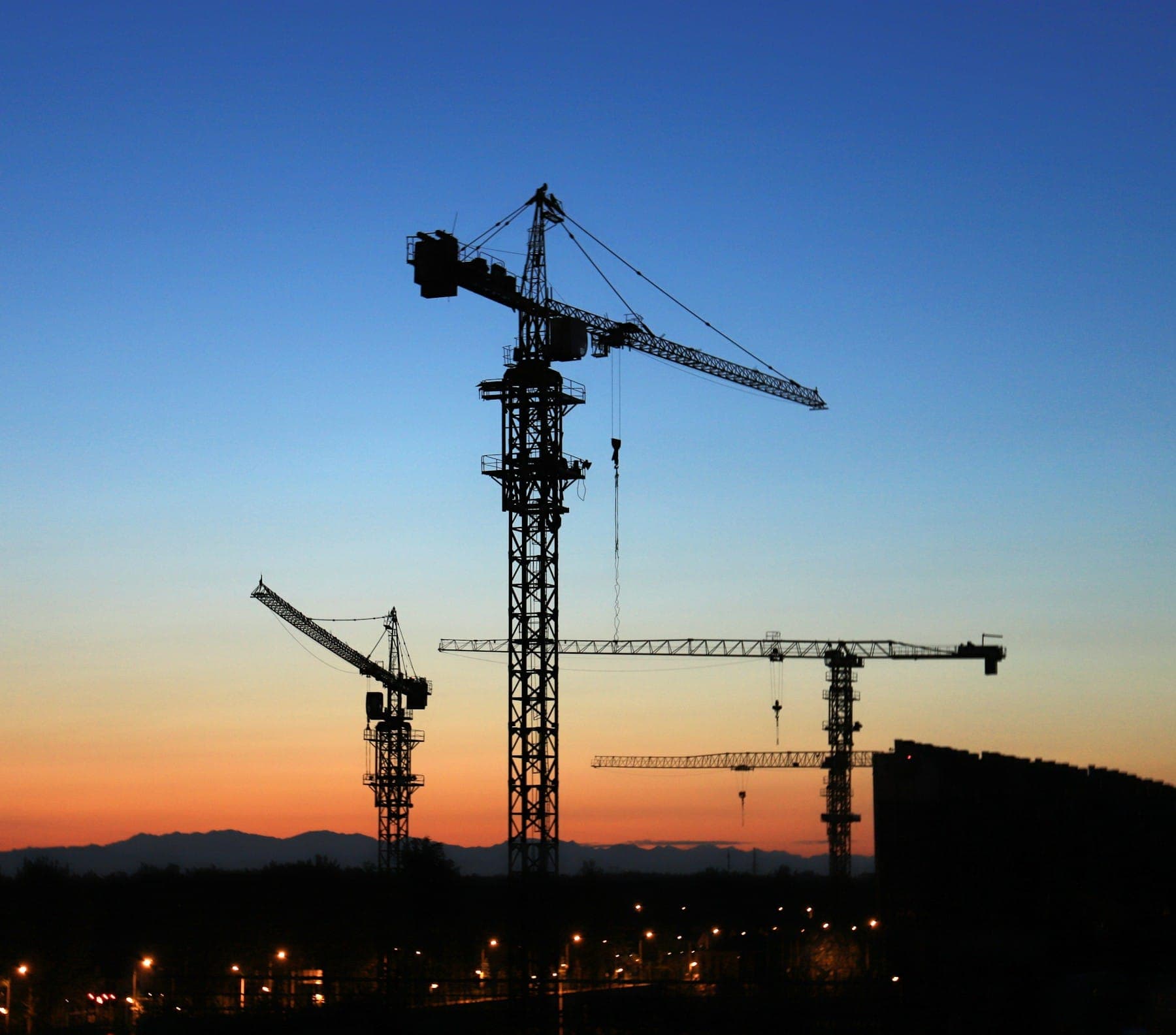 Tower cranes silhouetted against a sunset sky over a construction site