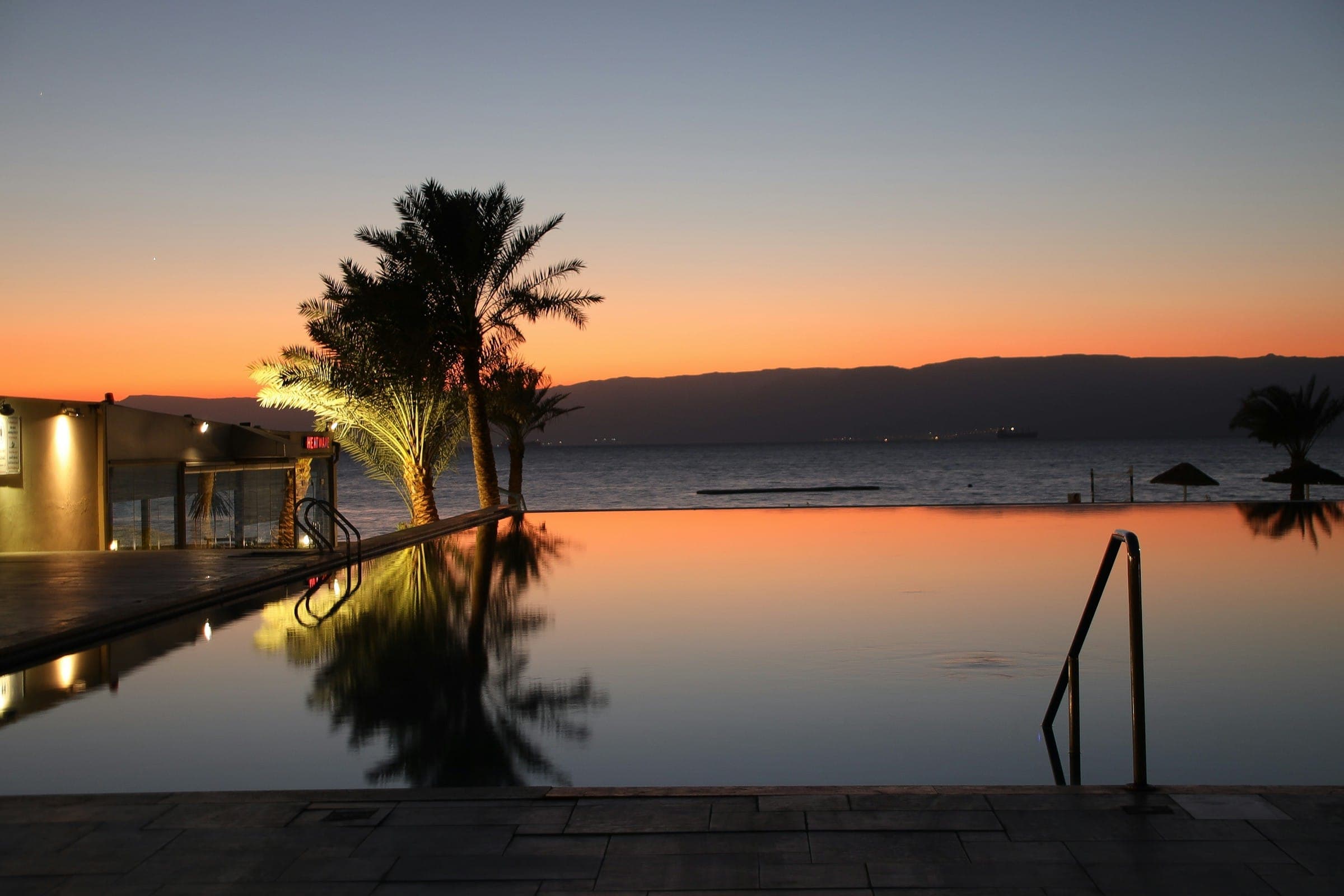 An infinity pool reflecting a palm tree and an orange sunset over the sea